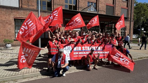 Gruppe von etwa 25 Personen in roten Warnwesten steht vor einem roten Backsteingebäude und hält mehrere rote Fahnen mit dem Aufdruck „EVG – Eisenbahn- und Verkehrsgewerkschaft“. In der Mitte wird ein rotes Banner gehalten mit dem gut lesbaren Text „WIR STREIKEN!“ sowie darunter „KEIN ABSCHLUSS OHNE GEWERKSCHAFT“. Einige Personen stehen, andere knien vor dem Banner. Sonniges Tageslicht, gepflasterter Vorplatz und Straße im Vordergrund.
