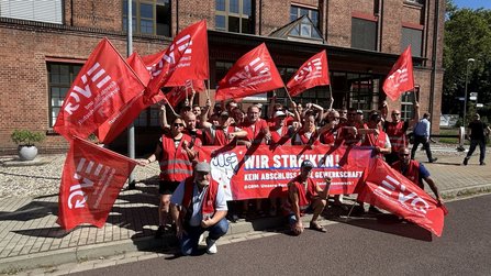 Gruppe von etwa 25 Personen in roten Warnwesten steht vor einem roten Backsteingebäude und hält mehrere rote Fahnen mit dem Aufdruck „EVG – Eisenbahn- und Verkehrsgewerkschaft“. In der Mitte wird ein rotes Banner gehalten mit dem gut lesbaren Text „WIR STREIKEN!“ sowie darunter „KEIN ABSCHLUSS OHNE GEWERKSCHAFT“. Einige Personen stehen, andere knien vor dem Banner. Sonniges Tageslicht, gepflasterter Vorplatz und Straße im Vordergrund.