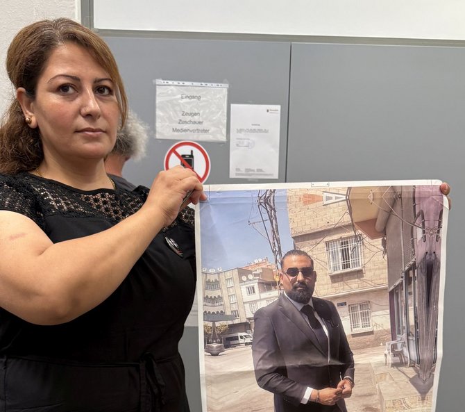 Frau hält ein großes Poster mit einem Foto eines Mannes im Anzug, der im Freien steht. Im Hintergrund sind eine Tür, Hinweisschilder und ein Piktogramm für ein Handyverbot zu sehen.