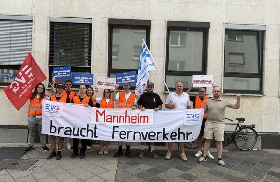 Gruppe von etwa zwölf Personen steht vor der Fassade eines Gebäudes auf dem Gehweg. Mehrere Personen tragen orangefarbene Warnwesten und halten Fahnen sowie Schilder hoch. Vor der Gruppe wird ein großes weißes Banner gehalten mit den Logos „EVG“ und dem Text „Mannheim braucht Fernverkehr“. Auf blauen Schildern steht „Damit Deutschland nicht den Anschluss verliert“, auf weißen Schildern „Schiene öffnen: Ja, Städte opfern: Nein!“. Links ist eine rote EVG-Fahne, in der Mitte eine weiße Fahne mit EVG-Logo zu sehen. Rechts neben der Gruppe steht ein Fahrrad.