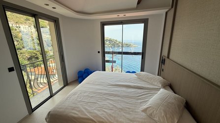 Helles Schlafzimmer mit Doppelbett vor großen Fenstern und Glastüren. Durch die Fenster sind Meer, Küstenhang und Boote in einer Bucht sichtbar. Neben dem Bett liegen mehrere blaue Ballons auf dem Boden. Ein schmaler Balkon mit Metallgeländer verläuft entlang der Fensterfront.