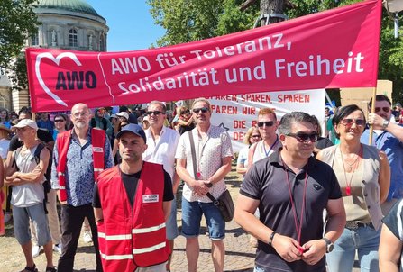 Gruppe von Menschen vor einem großen roten Banner mit der Aufschrift „AWO für Toleranz, Solidarität und Freiheit“. Mehrere Personen tragen rote Warnwesten. Hinter dem Banner stehen weitere Demonstrationsteilnehmende, Fahnen und Transparente. Im Hintergrund sind Bäume und ein rundes Gebäude mit Kuppeldach zu sehen.