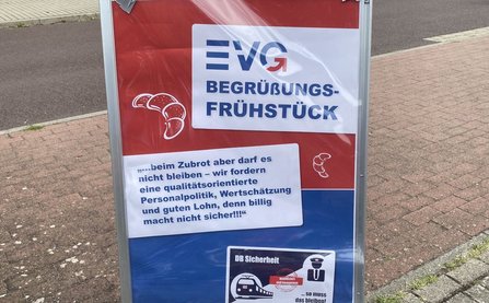 Freistehendes Werbeschild auf einem gepflasterten Gehweg. Oben steht groß „EVG BEGRÜSSUNGS-FRÜHSTÜCK“. Darunter der Text: „…beim Zubrot aber darf es nicht bleiben – wir fordern eine qualitätsorientierte Personalpolitik, Wertschätzung und guten Lohn, denn billig macht nicht sicher!!!“ Links und rechts sind Croissant-Symbole abgebildet. Im unteren Bereich befindet sich eine Grafik mit der Überschrift „DB Sicherheit“.