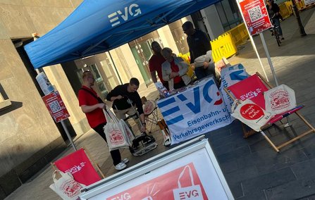 Informationsstand unter einem blauen „EVG“-Pavillon in einer Fußgängerpassage. Auf dem Tisch liegen Broschüren und Werbematerial. Mehrere Personen befinden sich am Stand; eine Person hält mehrere Stofftaschen. Im Vordergrund steht ein Werbeaufsteller mit rotem Plakat. Rechts ist ein Plakat mit QR-Code sichtbar, im Hintergrund gelbe Absperrelemente und Gebäudefassaden.
