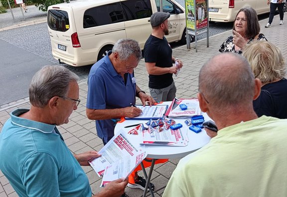 Mehrere Personen stehen an einem kleinen runden Stehtisch auf einem Gehweg und lesen sowie unterschreiben Formulare; auf dem Tisch liegen Flyer, Kugelschreiber und kleine Werbeartikel, im Hintergrund stehen Fahrzeuge und Passanten.