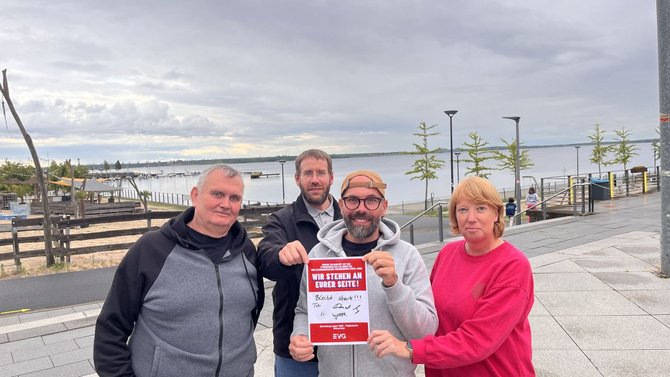 Vier Personen stehen auf einer Uferpromenade vor einem Gewässer. Die Person in der Mitte hält ein rot-weißes Schild mit dem deutlich lesbaren Text „WIR STEHEN AN EURER SEITE!“. Darunter sind handschriftliche Notizen und Unterschriften zu sehen. Im Hintergrund befinden sich ein Steg mit Bootsanlegern, mehrere junge Bäume, Geländer, Laternen und ein bewölkter Himmel.
