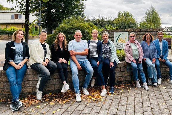 Neun Personen sitzen nebeneinander auf einer niedrigen Steinmauer vor dem Eingangsbereich der Vital-Klinik Dreizehnlinden im Freien. Die Gruppe ist auf einer gepflasterten Fläche vor einer Steinmauer versammelt. Hinter den Personen steht ein Schild mit der Aufschrift „Vital-Klinik Dreizehnlinden“. Die meisten Personen tragen Freizeitkleidung in gedeckten Farben. Einige sitzen direkt auf der Mauer, andere lehnen sich leicht an. Vor der Mauer liegen verstreut herbstliche Blätter auf dem Boden.