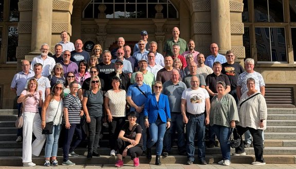 Gruppenfoto auf einer breiten Steintreppe vor einem Gebäude mit großen Säulen und einem Eingangsportal. Mehr als vierzig Personen stehen oder sitzen auf den Stufen in mehreren Reihen. Das Gebäude zeigt eine symmetrische Fassade mit Fenstern und verzierten Steinflächen im Hintergrund.