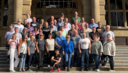 Gruppenfoto auf einer breiten Steintreppe vor einem Gebäude mit großen Säulen und einem Eingangsportal. Mehr als vierzig Personen stehen oder sitzen auf den Stufen in mehreren Reihen. Das Gebäude zeigt eine symmetrische Fassade mit Fenstern und verzierten Steinflächen im Hintergrund.