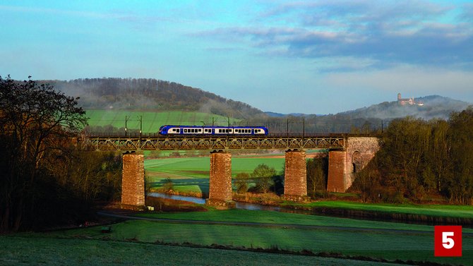 Blauer und weißer Triebwagen auf einer hohen Eisenbahnbrücke mit mehreren gemauerten Pfeilern über einem Fluss. Umgeben von Feldern, Bäumen und bewaldeten Hügeln. Rechts auf einem Hügel ist ein Gebäude sichtbar. Unten rechts befindet sich eine rote Markierung mit der Zahl „5“.