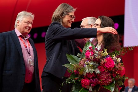 Mehrere Personen auf einer Bühne. Eine Person überreicht einer anderen Person einen großen Blumenstrauß mit roten und rosa Blumen sowie grünem Blattwerk. Weitere Personen stehen daneben. Der Hintergrund besteht aus einer roten Wand mit Bühnenbeleuchtung.
