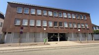 Dreigeschossiges Gebäude mit brauner, gekachelter Fassade und mehreren Reihen rechteckiger Fenster mit Jalousien; mittig ein gläserner Eingangsbereich. Davor Gehweg mit Pflastersteinen, zwei runde Außenlampen, ein grüner Mülleimer und ein Verkehrsschild mit Parkverbot. Verkehrsstraße im Vordergrund.