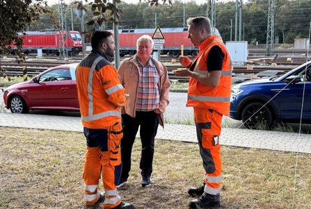 Drei Personen stehen auf einer Grasfläche neben Bahngleisen und unterhalten sich. Zwei Personen tragen orangefarbene Warnkleidung, eine Person trägt eine helle Jacke und ein kariertes Hemd. Im Hintergrund stehen rote Lokomotiven auf den Gleisen, daneben sind geparkte Autos und ein Warnschild sichtbar.