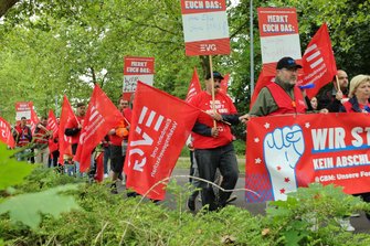 Demonstrationszug auf einem Weg zwischen Bäumen und Sträuchern. Zahlreiche rote EVG-Fahnen werden getragen. Im Vordergrund ist ein teilweise sichtbares Banner mit der Aufschrift „WIR STREIKEN!“ zu sehen. Mehrere Schilder mit der Überschrift „MERKT EUCH DAS:“ werden entlang des Zuges hochgehalten.