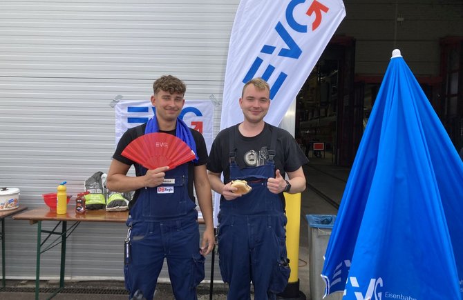 Zwei Personen in blauer Arbeitskleidung stehen vor einem Hallentor und einer weißen Fahne mit dem Schriftzug „EVG“. Die Person links hält einen roten Fächer, die Person rechts ein belegtes Brötchen und zeigt einen Daumen nach oben. Rechts im Vordergrund steht ein geschlossener blauer Sonnenschirm mit EVG-Aufdruck. Links im Hintergrund stehen ein Tisch mit Lebensmitteln und Grillzubehör sowie ein Topf.
