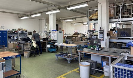 Innenraum einer Werkstatt mit Arbeitstischen, Maschinen, Werkzeugschränken und Regalen. Eine Person arbeitet an einem Arbeitsplatz im linken Bereich. Der Raum ist hell beleuchtet, mit weißen Wänden, Stützsäulen und Leuchtstofflampen an der Decke.