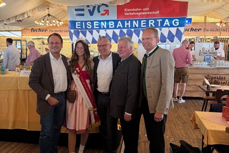 Fünf Personen stehen in einem Festzelt vor einem großen EVG-Banner mit der Aufschrift „Eisenbahnertag“ und posieren für ein gemeinsames Foto. Die Gruppe steht vor einem blau-weiß gestalteten Hintergrundbanner mit den Aufschriften „EVG“, „Eisenbahn- und Verkehrsgewerkschaft“ und „Eisenbahnertag“. Eine Person in traditioneller Festkleidung trägt eine Krone und eine Schärpe. Die übrigen vier Personen tragen überwiegend Anzug, Sakko oder traditionelle Festkleidung. Alle stehen dicht nebeneinander vor dem Banner.