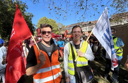 Zwei Personen stehen auf einer Grünfläche und halten Fahnen. Links ist eine rote Fahne mit EVG-Schriftzug zu sehen, rechts eine weiße Fahne mit teilweise sichtbarem Text, darunter „Frankfurt a.“ und „Betriebs“. Im Hintergrund befinden sich weitere Teilnehmende, Fahnen, Bäume und Gebäude.