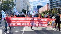 Demonstrationszug auf einer breiten Straße zwischen mehrstöckigen Gebäuden. Vorne wird ein großes rotes Banner getragen mit dem Text: „800 MILLIARDEN FÜR DIE BILDUNG“, darunter „mehr Kitas, kleinere Schulklassen, mehr Personal“ und „STATT FÜR KRIEGE, TOD UND PROFITE“. Hinter dem Banner gehen zahlreiche Personen mit weißen, roten und blau-weißen Fahnen.