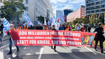 Demonstrationszug auf einer breiten Straße zwischen mehrstöckigen Gebäuden. Vorne wird ein großes rotes Banner getragen mit dem Text: „800 MILLIARDEN FÜR DIE BILDUNG“, darunter „mehr Kitas, kleinere Schulklassen, mehr Personal“ und „STATT FÜR KRIEGE, TOD UND PROFITE“. Hinter dem Banner gehen zahlreiche Personen mit weißen, roten und blau-weißen Fahnen.