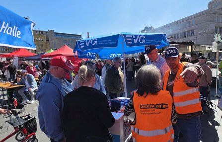 Belebter Stadtplatz mit vielen Menschen an Stehtischen und Informationsständen. In der Mitte steht ein blauer Sonnenschirm mit der Aufschrift „EVG Eisenbahn- und Verkehrsgewerkschaft“. Mehrere rote Zelte und weitere blaue Schirme sind sichtbar. Im Vordergrund tragen zwei Personen orangefarbene Warnwesten mit dem Text „GEMEINSAM GEHT MEHR“ und „Eisenbahn- und Verkehrsgewerkschaft“. Weitere sichtbare Texte sind „EVG“ auf Westen und Schirmen sowie „Halle“ auf einer blauen Fahne. Im Hintergrund stehen größere Gebäude und ein klarer blauer Himmel.