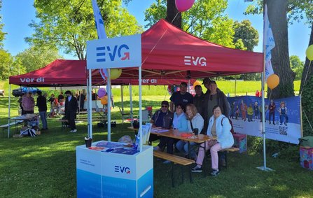 Mehrere rote Pavillons auf einer Grünfläche mit Tischen und Bänken darunter. Auf den Pavillons und Schildern ist „EVG“ sowie „ver.di“ zu lesen. Eine Gruppe von Personen sitzt und steht an einem Tisch; auf dem Tisch liegen Flyer und Materialien. Im Hintergrund sind weitere Pavillons, Bäume und Luftballons zu sehen; Tageslicht bei klarem Himmel.