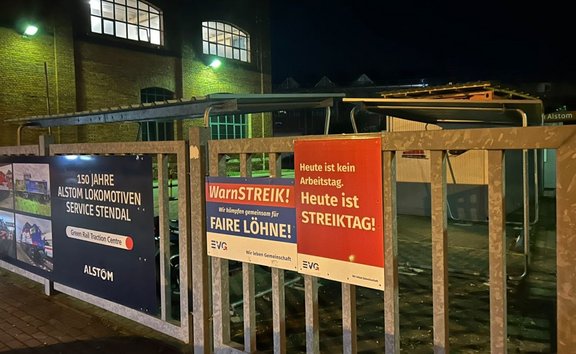 Nächtliche Aufnahme eines Metallzauns vor einem Industriegebäude mit beleuchteten Fenstern. Am Zaun hängen drei Schilder: links ein Alstom-Schild mit der Aufschrift „150 JAHRE ALSTOM LOKOMOTIVEN SERVICE STENDAL“, „Green Rail Traction Centre“ und „ALSTOM“ sowie mehreren Zugbildern; in der Mitte ein Schild mit „WarnSTREIK!“, „Wir kämpfen gemeinsam für FAIRE LÖHNE!“ und dem EVG-Logo; rechts ein rotes Schild mit der Aufschrift „Heute ist kein Arbeitstag. Heute ist STREIKTAG!“. Im Hintergrund sind überdachte Fahrradständer und weitere Betriebsgebäude sichtbar.