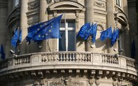 Mehrere blaue Flaggen mit gelben Sternen der Europäischen Union sind an schräg angebrachten Fahnenstangen an der Fassade eines hellen, verzierten Gebäudes befestigt. Vor den Fenstern befindet sich ein halbrunder Balkon mit steinernem Geländer und dekorativen Reliefs. Die Flaggen wehen nach außen, einige überlappen sich, während das Gebäude mit Säulen, Ornamenten und Fenstereinfassungen im Hintergrund zu sehen ist.