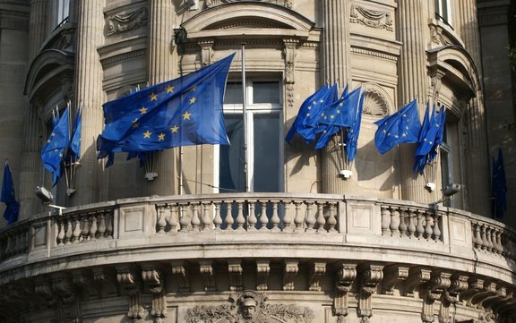 Mehrere blaue Flaggen mit gelben Sternen der Europäischen Union sind an schräg angebrachten Fahnenstangen an der Fassade eines hellen, verzierten Gebäudes befestigt. Vor den Fenstern befindet sich ein halbrunder Balkon mit steinernem Geländer und dekorativen Reliefs. Die Flaggen wehen nach außen, einige überlappen sich, während das Gebäude mit Säulen, Ornamenten und Fenstereinfassungen im Hintergrund zu sehen ist.