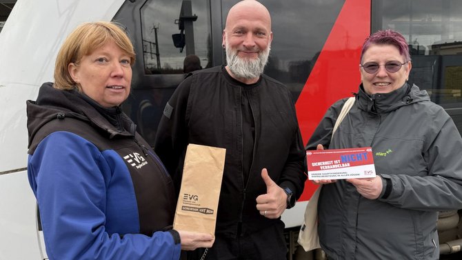 Drei Personen stehen vor einem Zug. Links trägt eine Person eine blaue Jacke mit schwarzer EVG‑Weste und hält eine braune Papiertüte mit EVG‑Aufdruck. In der Mitte steht eine Person in schwarzer Kleidung und zeigt mit der rechten Hand einen erhobenen Daumen. Rechts trägt eine Person eine graue Jacke und hält einen roten und weißen Flyer mit der Aufschrift „Schweigen ist nicht vertretbar“. Im Hintergrund sind die Außenwand und Fenster des Zuges zu sehen.