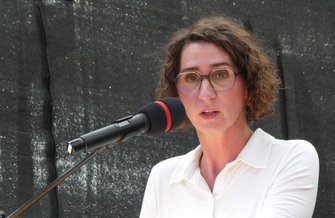 Person mit kurz gelockten Haaren steht vor einem Mikrofon mit rotem Windschutz. Die Person trägt ein weißes Oberteil. Hintergrund ist eine dunkle, strukturierte Fläche.