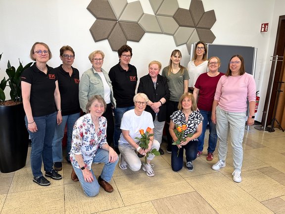 Eine Gruppe von zwölf Personen steht und kniet für ein Gruppenfoto in einem Innenraum; drei Personen im Vordergrund halten jeweils orangefarbene Blumen. Das Foto zeigt eine Gruppe von Personen, die sich in zwei Reihen angeordnet haben: neun Personen stehen im Hintergrund, drei Personen knien oder sitzen vorne. Zwei der vorderen Personen halten jeweils eine Rose mit orangenen Blüten, eine weitere Person vorne hält ebenfalls Blumen. Mehrere Personen im Hintergrund tragen schwarze Oberteile mit einem rot-weißen Logo. Die Gruppe befindet sich in einem hellen Raum mit beigefarbenem Boden und weißen Wänden. An der Wand hinter der Gruppe sind dekorative, sechseckige Elemente in verschiedenen Grau- und Beigetönen angebracht. Rechts im Bild sind eine Tür und ein Feuerlöscher zu sehen, links steht eine große Pflanze in einem schwarzen Topf.