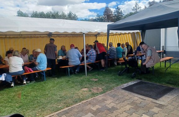 Mehrere Personen sitzen an Bierzeltgarnituren unter einem großen weiß-gelben Festzelt auf einer Rasenfläche. Rechts steht ein weiteres dunkles Pavillonzelt mit zusätzlichen Sitzplätzen. Im Vordergrund sind Rasen, ein gepflasterter Weg und eine schwarze Bodenmatte zu sehen. Im Hintergrund befinden sich Bäume und ein Gebäude mit heller Fassade. Der Himmel ist teilweise bewölkt.