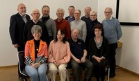 Gruppenfoto in einem Innenraum mit weißer Wand und hellem Holzboden. Insgesamt zwölf Personen sind zu sehen: vier Personen sitzen in der vorderen Reihe auf Stühlen, acht Personen stehen dicht dahinter. Die Gesichter aller Personen sind unkenntlich gemacht. Die sitzenden Personen tragen unter anderem eine orange Jacke, einen rosafarbenen Pullover, ein dunkles Kurzarmoberteil und eine dunkle Strickjacke. In der stehenden Reihe sind verschiedene Oberteile in dunklen und hellen Farben zu sehen, darunter Grau, Blau, Schwarz und ein rostfarbener Pullover. Rechts im Hintergrund ist der Rand einer Projektionsleinwand erkennbar.