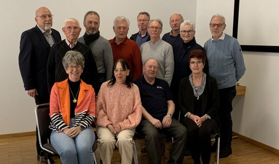 Gruppenfoto in einem Innenraum mit weißer Wand und hellem Holzboden. Insgesamt zwölf Personen sind zu sehen: vier Personen sitzen in der vorderen Reihe auf Stühlen, acht Personen stehen dicht dahinter. Die Gesichter aller Personen sind unkenntlich gemacht. Die sitzenden Personen tragen unter anderem eine orange Jacke, einen rosafarbenen Pullover, ein dunkles Kurzarmoberteil und eine dunkle Strickjacke. In der stehenden Reihe sind verschiedene Oberteile in dunklen und hellen Farben zu sehen, darunter Grau, Blau, Schwarz und ein rostfarbener Pullover. Rechts im Hintergrund ist der Rand einer Projektionsleinwand erkennbar.