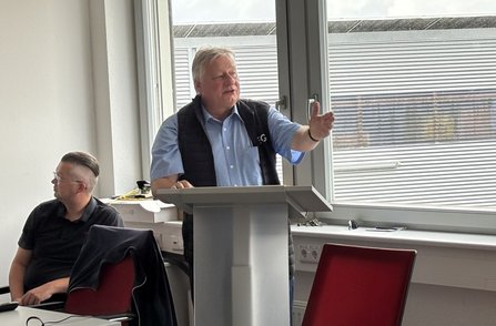 Eine Person steht an einem Rednerpult in einem Besprechungsraum und gestikuliert mit einer Hand. Neben dem Pult sitzt eine weitere Person an einem Tisch. Im Hintergrund befinden sich große Fenster, Stühle und technische Anschlüsse an der Wand.