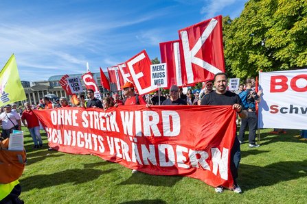 Gruppe von Demonstrierenden auf einer Rasenfläche mit roten Fahnen und einem großen roten Transparent. Auf dem Transparent steht: „OHNE STREIK WIRD SICH NICHTS VERÄNDERN“. Mehrere Schilder im Hintergrund tragen den Text „MERZ MUSS WEG“ und ein Schild zeigt „MERZ LECK EIER“. Im Hintergrund sind weitere Personen, Fahnen, Bäume und Gebäude zu sehen.
