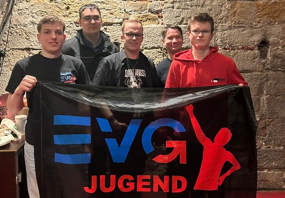 Fünf Personen stehen dicht beieinander vor einer groben Steinwand und halten ein großes schwarzes Banner mit der Aufschrift „EVG JUGEND“, auf dem blaue, rote und weiße grafische Elemente zu sehen sind.