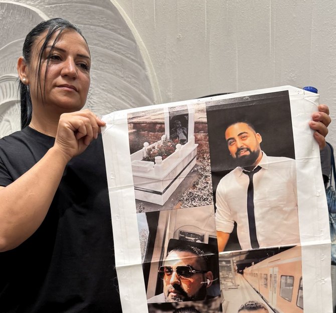 Frau hält ein großes Poster mit mehreren Fotos eines Mannes sowie einem Bild eines Grabes. Sie steht vor einer hellen Wand und blickt leicht zur Seite. Das Poster zeigt mehrere Porträts desselben Mannes in unterschiedlichen Situationen.