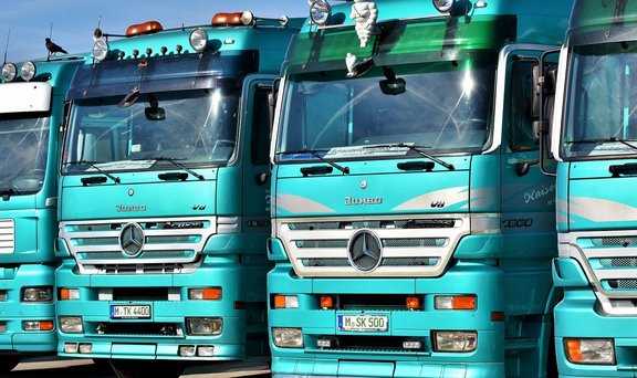 Frontansicht mehrerer türkisfarbener Lastwagen, dicht nebeneinander geparkt. Sichtbar sind die Kühlergrills mit Mercedes‑Stern, Frontscheinwerfer, Zusatzleuchten auf den Dächern und teilweise lesbare Kennzeichen. Auf einer Windschutzscheibe steht „Schaustellerfahrzeug“. Der Himmel ist klar und blau.