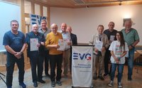 Gruppenfoto in einem Veranstaltungsraum mit Holzdecke und hellem Boden. Zehn Personen stehen vor einem Rednerpult mit einem weißen Tuch, auf dem das blaue Logo „EVG“ sowie der Text „Eisenbahn- und Verkehrsgewerkschaft“ und „www.evg-online.org“ zu sehen sind. Mehrere Personen halten Urkunden oder Broschüren in den Händen. Im Hintergrund steht links ein Banner mit dem EVG-Logo. An den Wänden befinden sich Leuchten, Türen und Fenster.