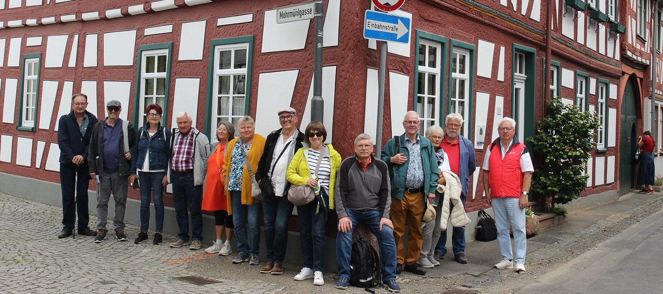 Eine Gruppe von etwa zwölf Personen steht an einer Straßenecke vor einem Fachwerkhaus und blickt zur Kamera. Die Gruppe steht auf einem gepflasterten Gehweg direkt an der Ecke eines rot-weißen Fachwerkgebäudes. Einige Personen lehnen an der Hauswand, eine Person sitzt auf einem niedrigen Vorsprung. Mehrere tragen Jacken, Hemden oder Westen in verschiedenen Farben, darunter Rot, Gelb, Blau und Orange. Taschen, Rucksäcke und Kopfbedeckungen sind ebenfalls zu sehen. Rechts vorne trägt eine Person eine rote Weste mit der Aufschrift „EVG“.