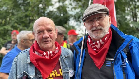 Zwei Personen im Vordergrund tragen rote Halstücher. Eine Person trägt eine Jeansjacke, die andere eine blaue Jacke und eine Schiebermütze. Im Hintergrund stehen weitere Personen, einige mit roten Kappen und Fahnen, vor Bäumen.