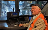 Person mit orangefarbener Warnweste und dunkler Kappe sitzt an einem Bedienpult in einem Schienenfahrzeug oder Simulator. Vor der Frontscheibe sind industrielle Anlagen, Metallkonstruktionen und Gleisbereiche sichtbar. Mehrere Bildschirme und Bedienelemente befinden sich auf dem Steuerstand.