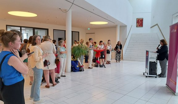 Mehrere Personen stehen in einem großen, hellen Raum und richten ihren Blick auf eine Frau, die an einem Rednerpult mit Mikrofon spricht. Neben ihr steht ein pinkfarbener DGB-Aufsteller. Im Raum befinden sich hohe Decken, eine Treppe, Pflanzen und Stehtische.