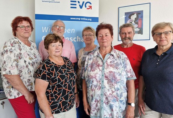 Sieben Personen stehen in zwei Reihen vor einem EVG-Roll-up-Banner. Auf dem Banner sind das EVG-Logo, die Internetadresse „www.evg-online.org“ und der Text „Wir leben Gemeinschaft“ sichtbar. An der Wand rechts neben dem Banner hängt ein gerahmtes Bild.