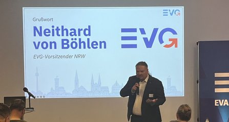 Eine Person spricht mit einem Handmikrofon vor einer Projektionsfläche. Auf der Leinwand steht „Grußwort Neithard von Böhlen“ sowie „EVG-Vorsitzender NRW“ neben dem EVG-Logo. Im Vordergrund sind sitzende Teilnehmende von hinten zu sehen. Rechts steht ein blaues Banner mit der Aufschrift „EVA Akademie“.