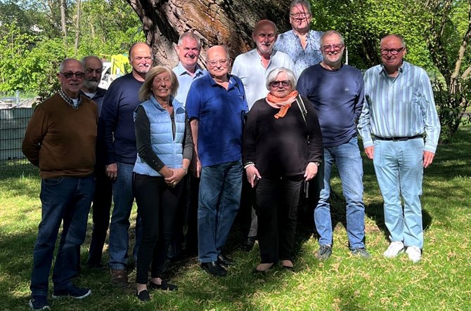 Gruppe von etwa zwölf Personen steht im Freien auf einer Rasenfläche vor einem großen Baum. Die Personen stehen dicht nebeneinander in zwei Reihen und tragen überwiegend langärmelige Oberteile, Westen, Jacken und Jeans. Im Hintergrund sind grüne Bäume, Sträucher und ein Metallzaun zu sehen. Das Licht ist helles Tageslicht, der Boden ist mit Gras bedeckt.