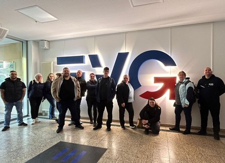Innenraum mit großer Wandgestaltung aus beleuchteten Buchstaben „EVG“ und einem Pfeilsymbol. Mehrere Personen stehen davor in einer Reihe; eine Person sitzt oder kniet im Vordergrund. Der Boden ist gefliest, links befindet sich ein Eingangsbereich mit Glasflächen.