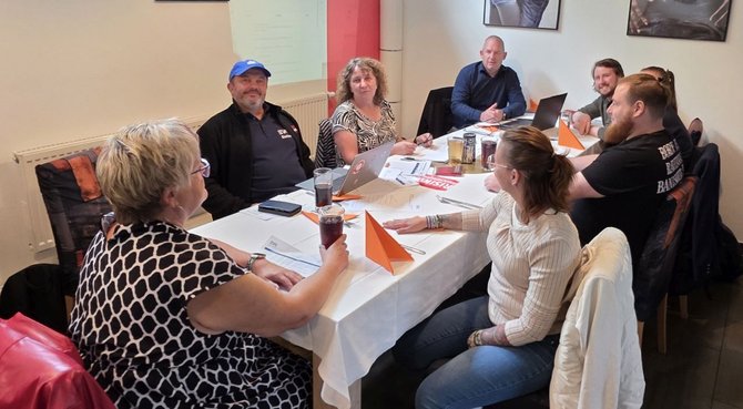 Acht Personen sitzen an einem rechteckigen Tisch in einem Besprechungsraum. Auf dem Tisch liegen Unterlagen, Namensschilder, Laptops, Gläser und Getränke. An der Stirnseite des Tisches steht ein geöffneter Laptop. An der Wand ist eine projizierte Präsentationsfolie zu sehen, darüber und daneben hängen gerahmte Bilder. Ein Teil der Personen blickt zur Präsentation, andere zum Tisch.